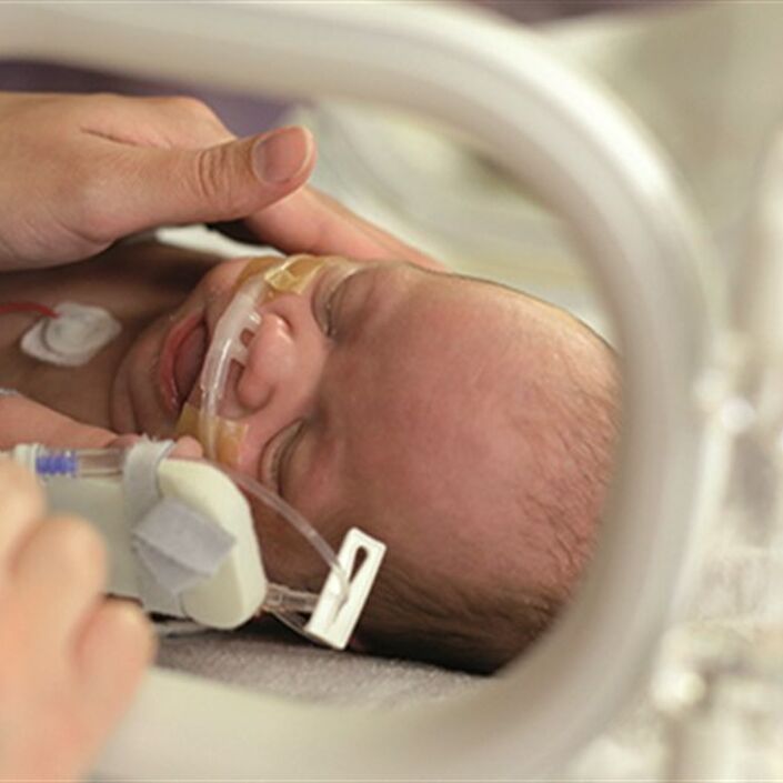 Baby in incubator with an adult's hand touching the baby's face