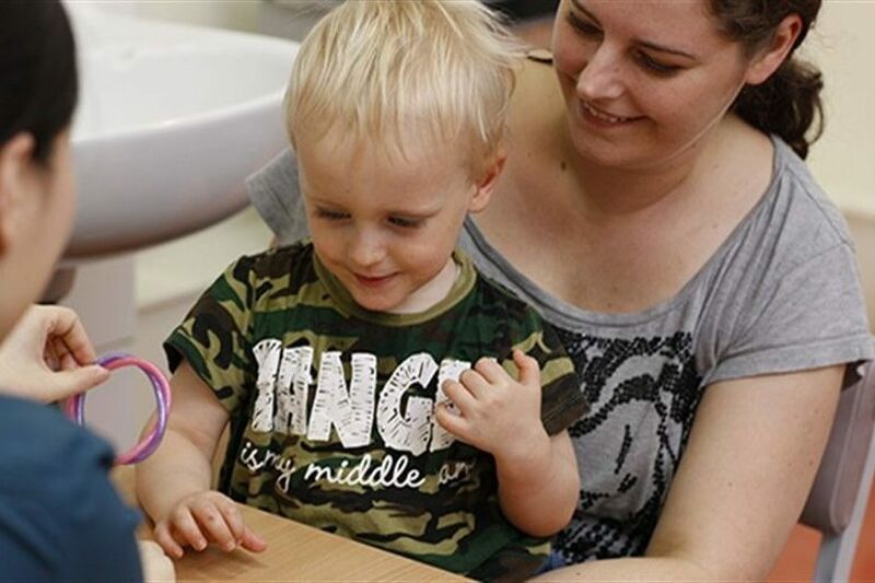 Little boy aged about 3 sitting on his mum at a table doing a task with health professional who has their back to camera