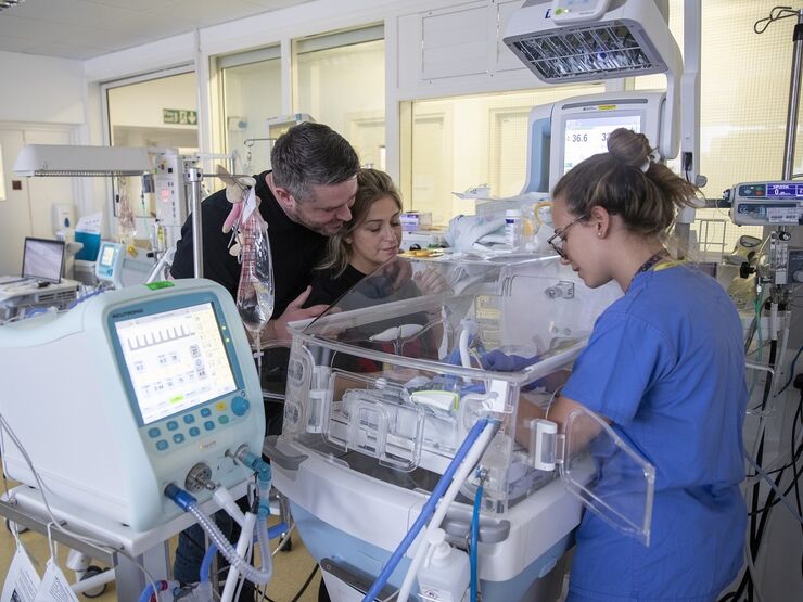 Parents with nurse in unit by cot looking at their baby while the nurse provides care for the baby