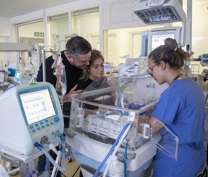 Parents with nurse in unit by cot looking at their baby while the nurse provides care for the baby
