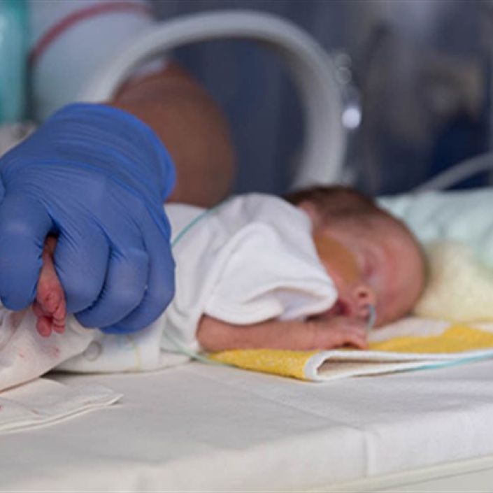 Close up of baby in cot lying on front with health professional wearing gloves touching the baby's feet