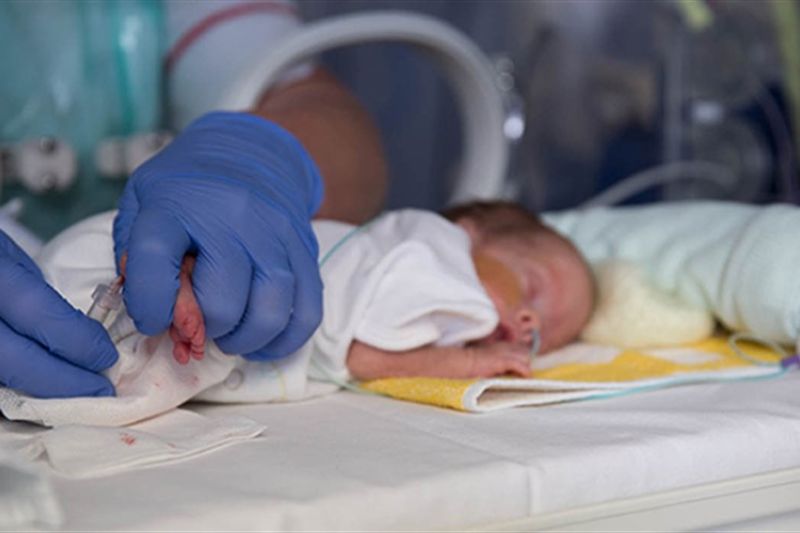 Close up of baby in cot lying on front with health professional wearing gloves touching the baby's feet