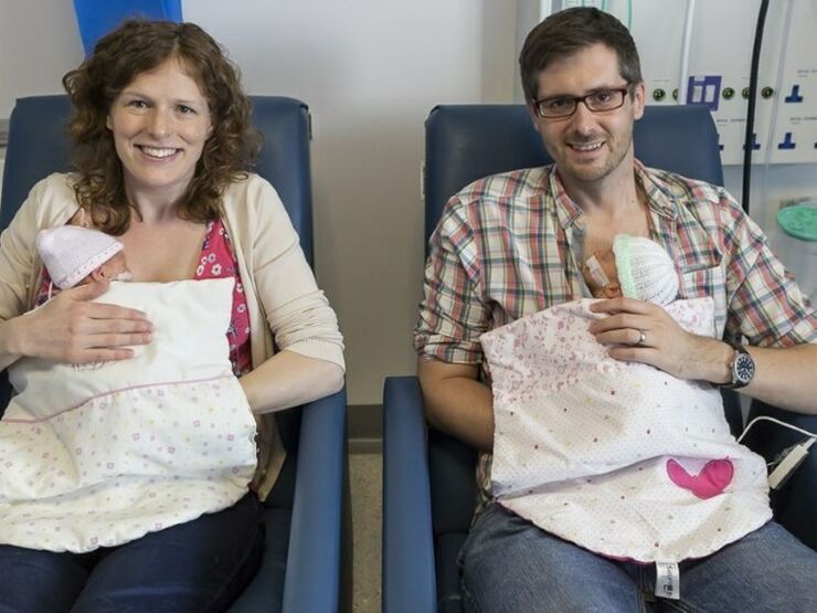 Mum and dad sitting in hospital chairs holding a baby twin each smiling at the camera