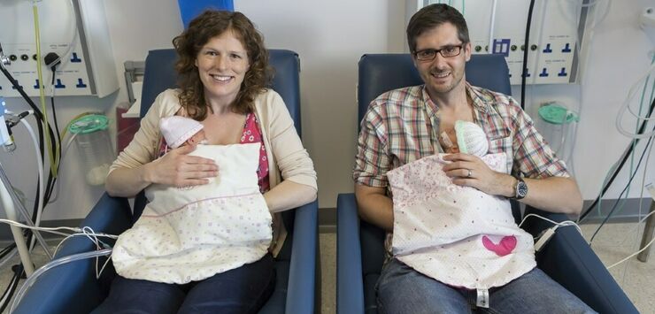 Mum and dad sitting in hospital chairs holding a baby twin each smiling at the camera