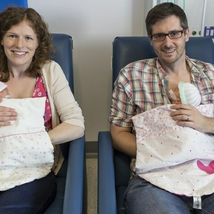Mum and dad sitting in hospital chairs holding a baby twin each smiling at the camera