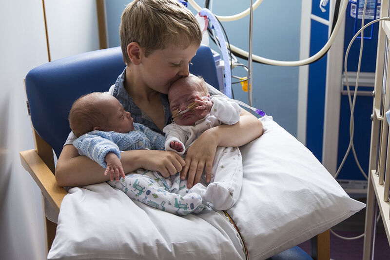 Twin babies being held by an older brother sitting on hospital chair with babies on a pillow and the older brother kissing one of their heads
