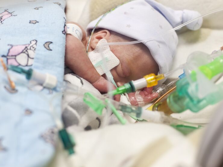Baby asleep covered in blanket and wearing a hat attached to several tubes