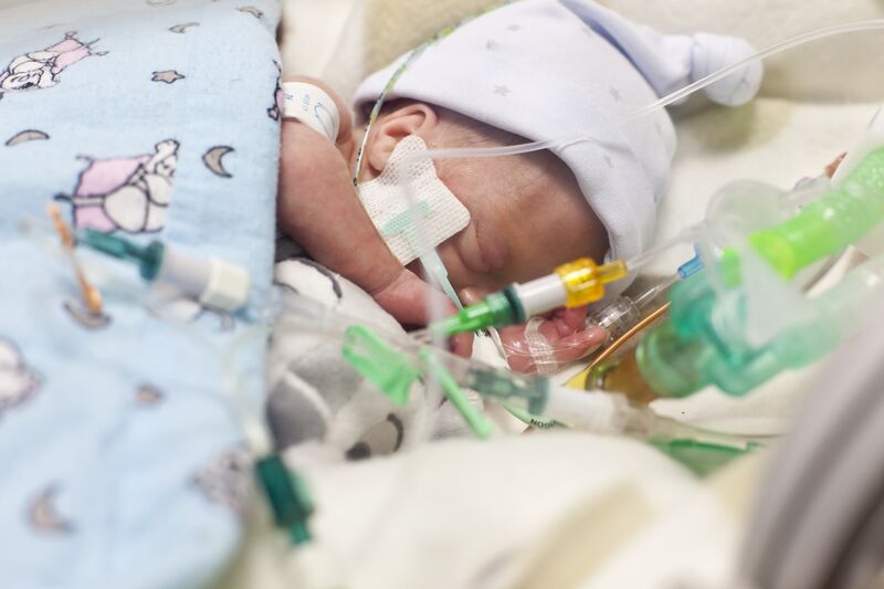 Baby asleep covered in blanket and wearing a hat attached to several tubes