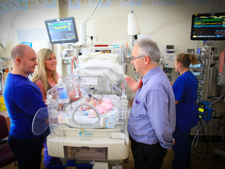 Parents standing on one side of an incubator with their baby inside talking to a doctor on the other side of the incubator