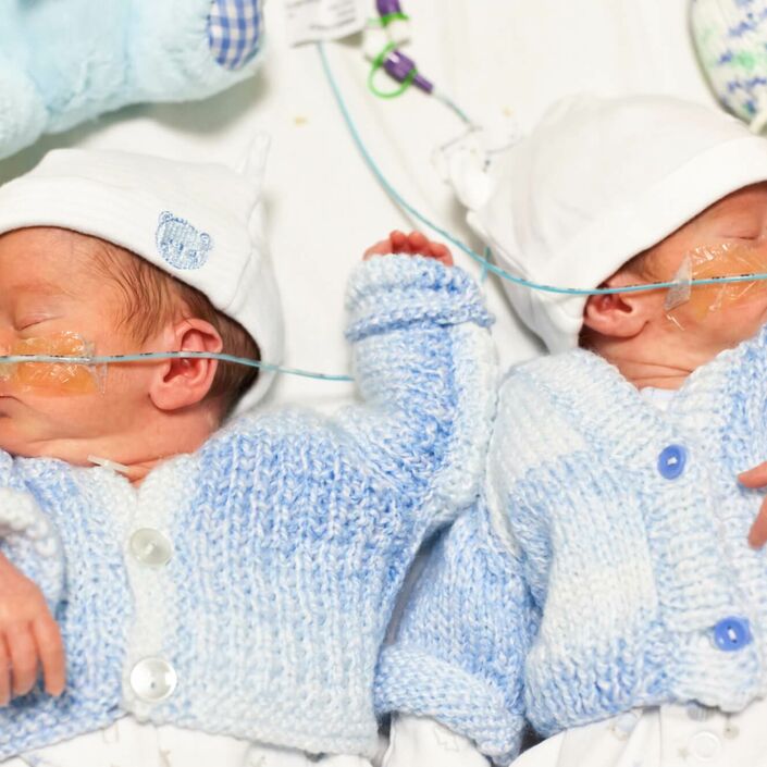 Twin babies asleep in hospital bed wearing blue cardigans and little hats with teddies above them