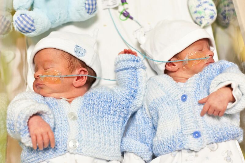 Twin babies asleep in hospital bed wearing blue cardigans and little hats with teddies above them
