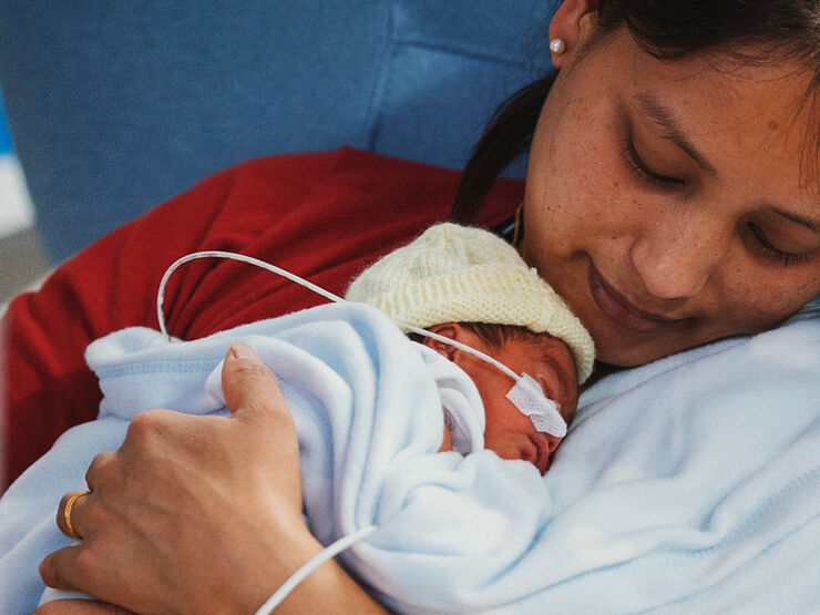 Mother holding and looking down at her newborn on her chest. Baby wearing wool hat and being tube-fed