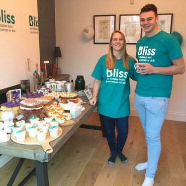 Two people in Bliss t-shirts standing next to their bake sale, with cakes on a table
