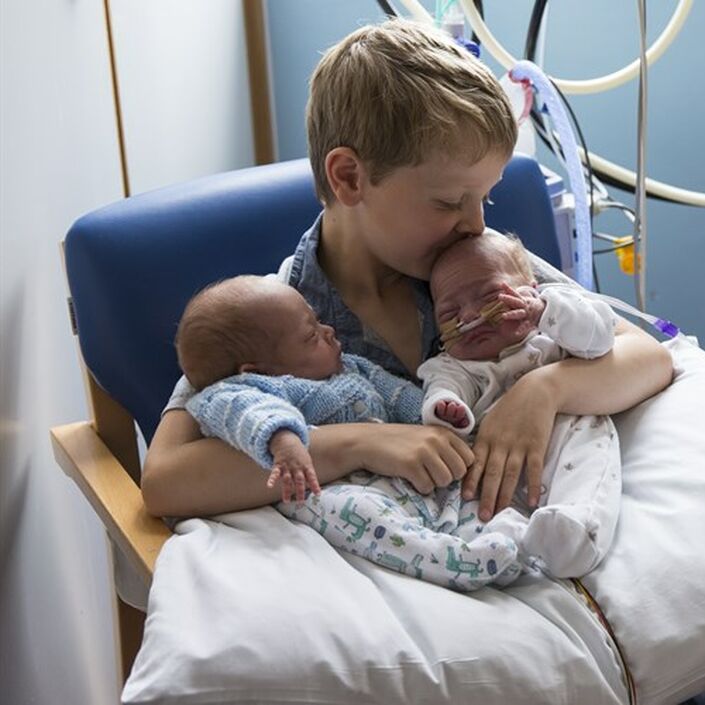 A boy holding his premature twin siblings in a hospital, kissing one on the head