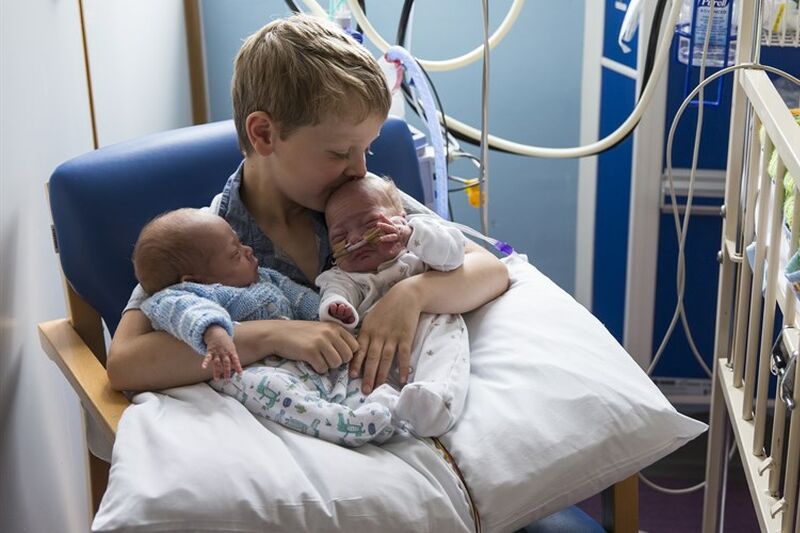 A boy holding his premature twin siblings in a hospital, kissing one on the head