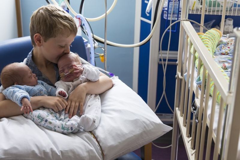 Brother holding twin siblings on his lap in hospital while kissing one on the head
