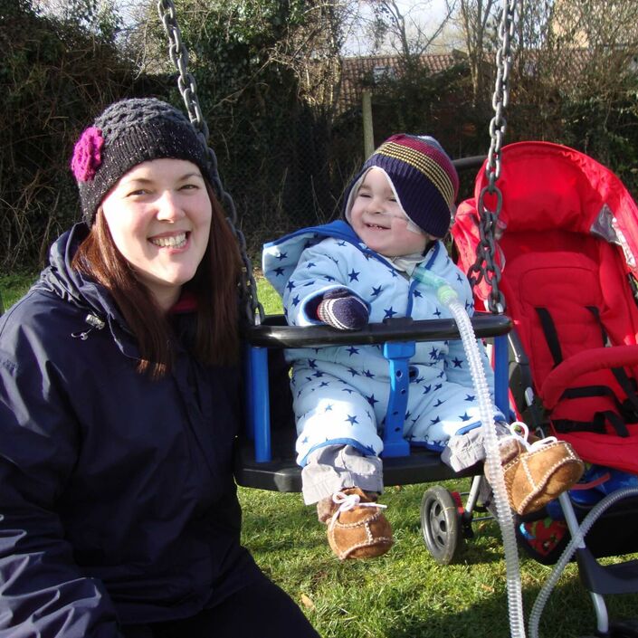 Mum smiling to the camera beside her baby on a swing attached to a breathing tube