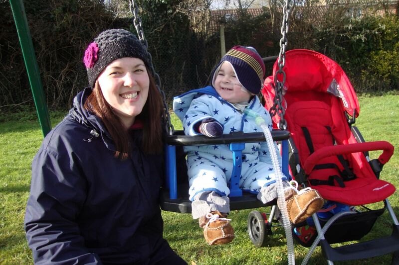 Mum smiling to the camera beside her baby on a swing attached to a breathing tube