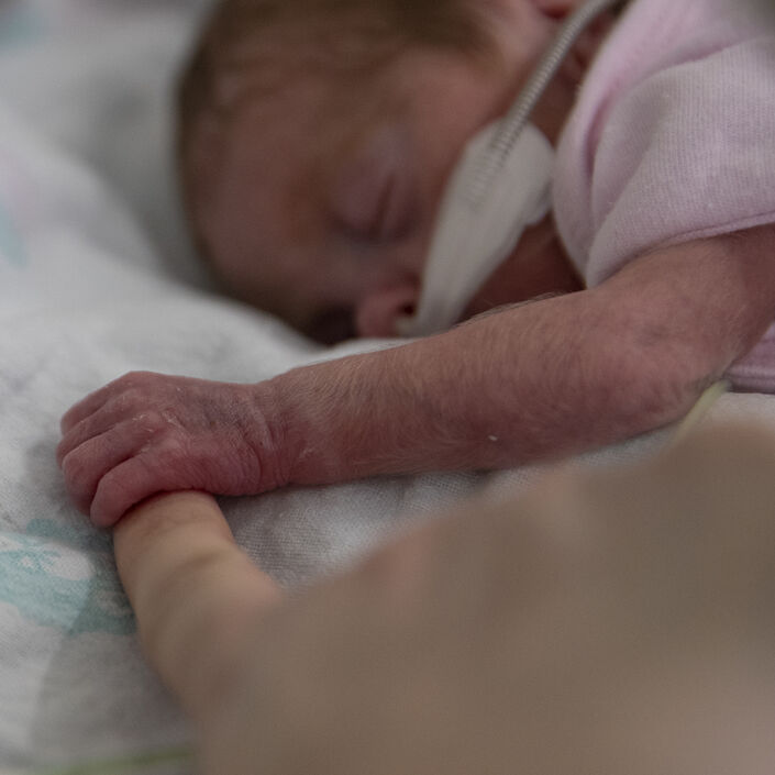 Baby lying on front asleep holding the finger of an adult