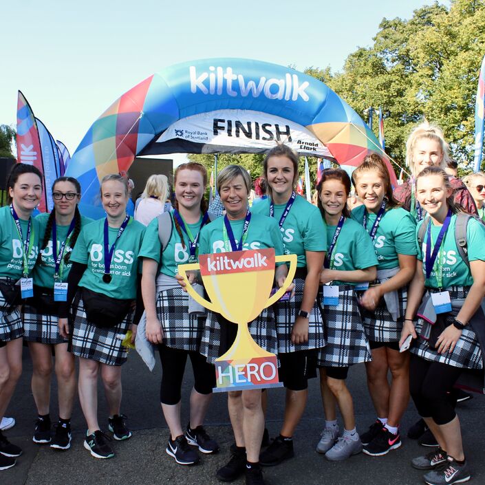 Group of women wearing kilts and bliss t-shirts at Kiltwalk finish line holding foamboard cut-out Kiltwalk hero trophy