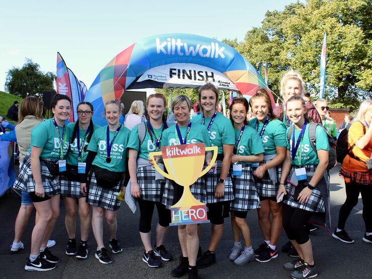 Group of women wearing kilts and bliss t-shirts at Kiltwalk finish line holding foamboard cut-out Kiltwalk hero trophy