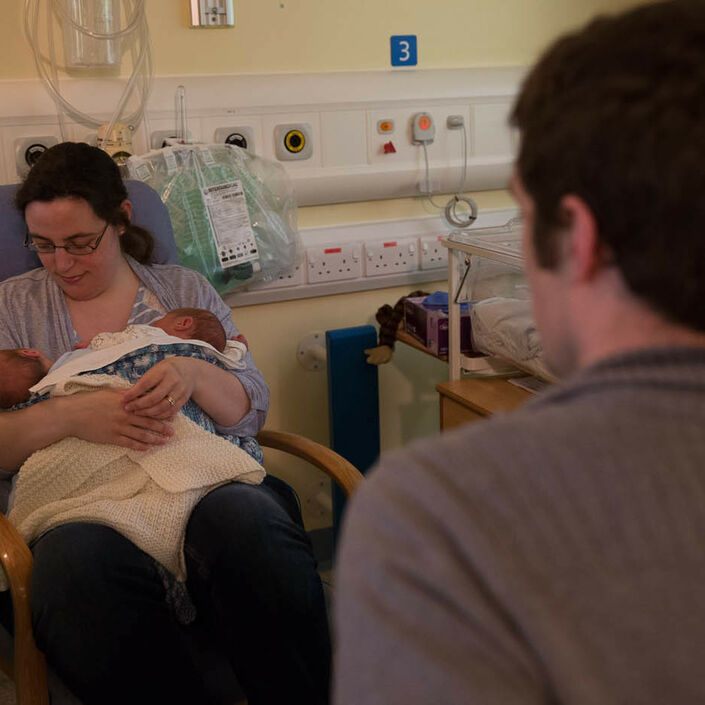 Dad in foreground looking away from camera towards Mum who is sitting holding and looking at her twins in her arms