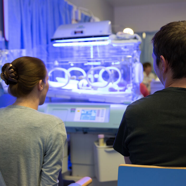 Mum and Dad on the neonatal unit sat down with their backs to the camera, looking towards an incubator and blue light from phototherapy treatment.