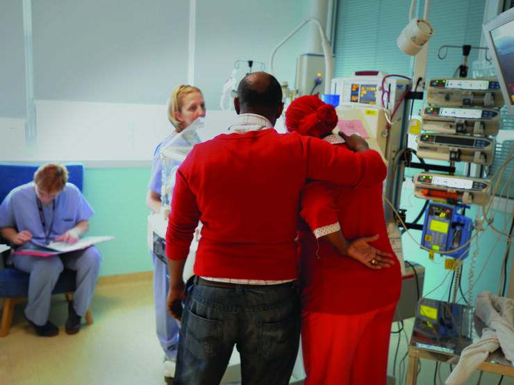 Facing away from the camera, Dad puts arm around Mum in a caring way whilst looking at baby in an incubator surrounded by two health professionals