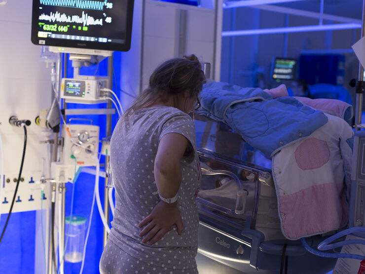 Mum standing by incubator in low light, looking at her baby and away from the camera