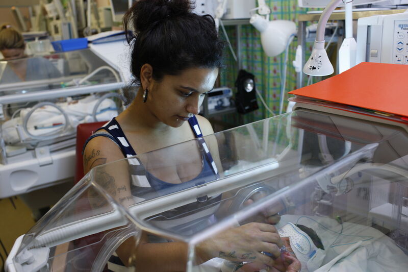 Mum standing looking into incubator at her baby who is wearing an eye mask. She is reaching through the incubator to comfort her baby.