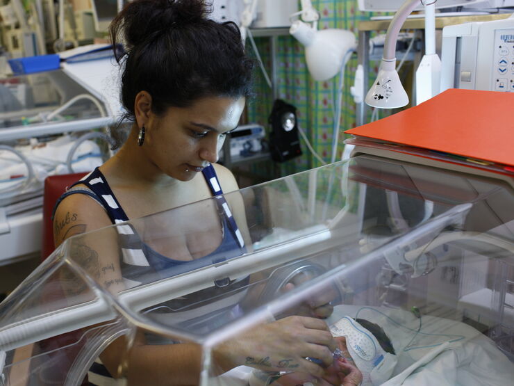 Mum standing looking into incubator at her baby who is wearing an eye mask. She is reaching through the incubator to comfort her baby.