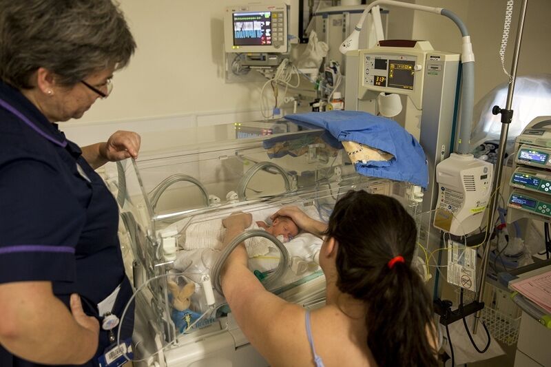 Mum sitting down looking away from the camera reaching into the incubator with both hands, holding her baby and looking at eye level. A nurse is standing next to her looking into the incubator
