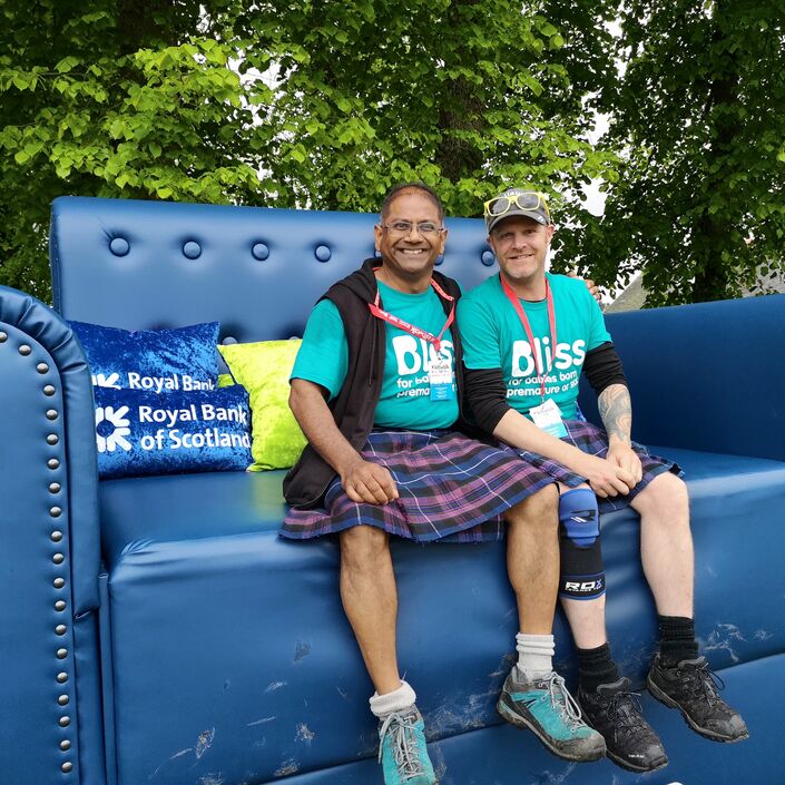 Two kiltwalk fundraisers wearing Bliss t-shirts and kilts sat on a extremely large sofa