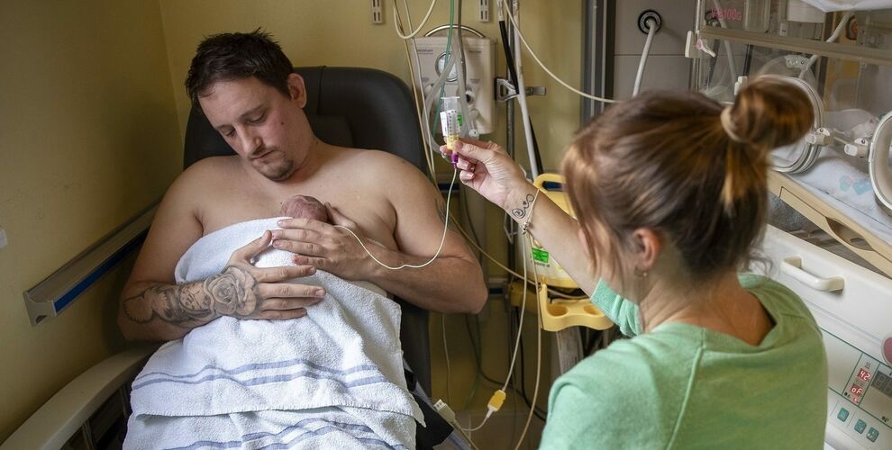 A dad is doing skin-to-skin, sitting in a hospital chair, while a mum is holding up the tube feeding their baby.