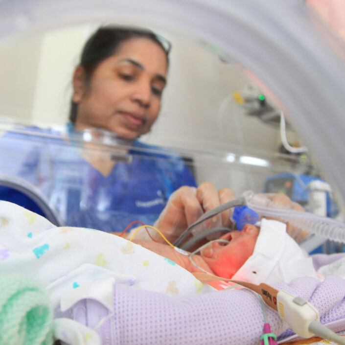 Baby in incubator on neonatal unit with nurse in background attending to the baby