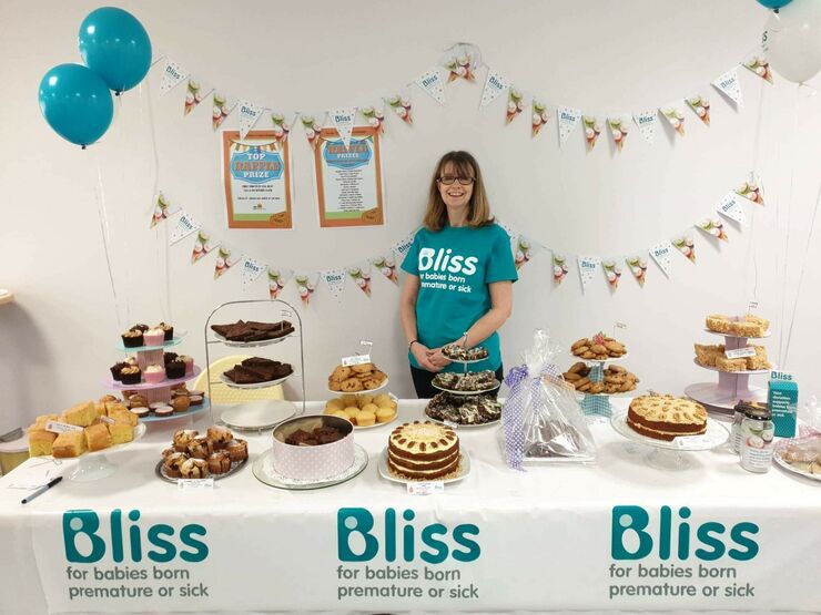 Lady wearing Bliss t-shirt standing behind a table of cakes and bunting and balloons behind, and table decorated with Bliss logo