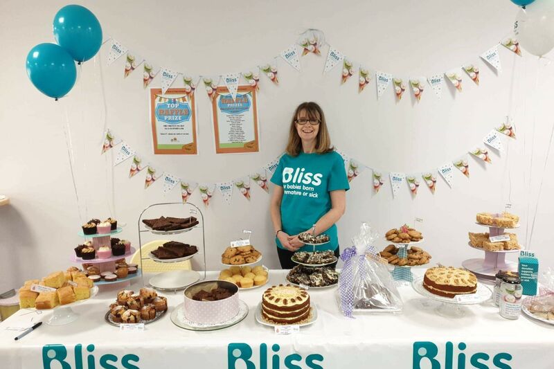Lady wearing Bliss t-shirt standing behind a table of cakes and bunting and balloons behind, and table decorated with Bliss logo