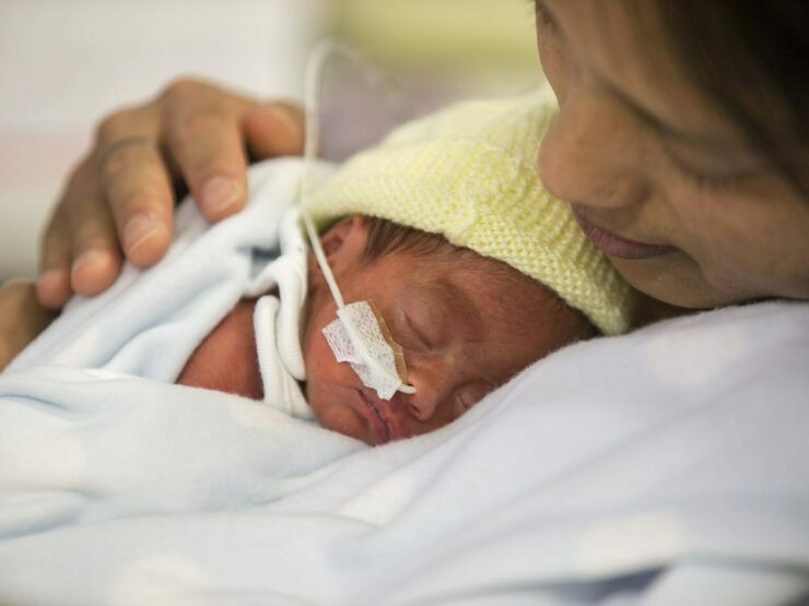 A mother leaning back with her baby sleeping on her chest. Baby is wearing a knitted, yellow bonnet and has a nasogastric tube.