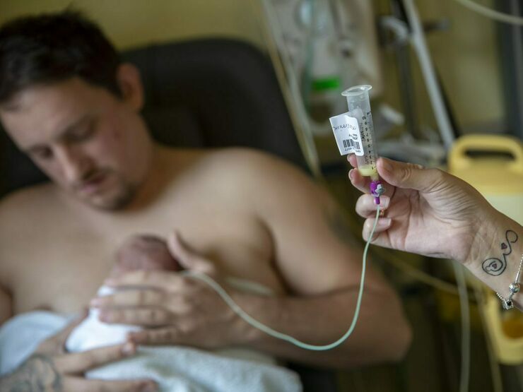 A father is sitting with his baby cuddling on his chest. The baby is receiving milk through a tube which is being held by his mother.