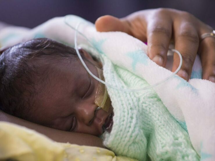A baby sleeps on his mother's chest, wrapped in blankets, with a nasogatric tube taped to his face