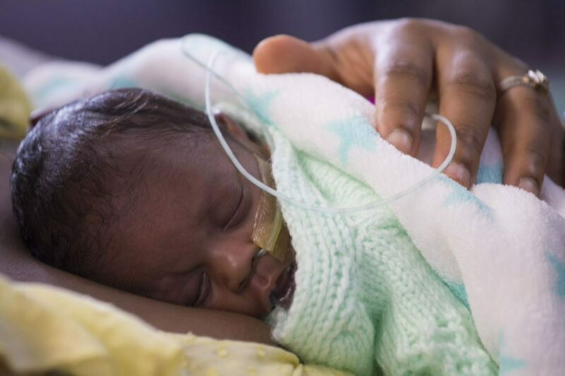 A baby sleeps on his mother's chest, wrapped in blankets, with a nasogatric tube taped to his face