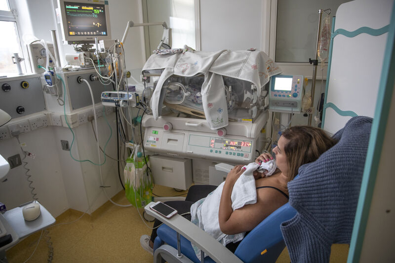 Mum sat on a hospital chair with her baby resting on her chest. In the background is an incubator and monitor
