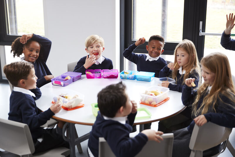 Group of primary school children sat at a round table having lunch and laughing