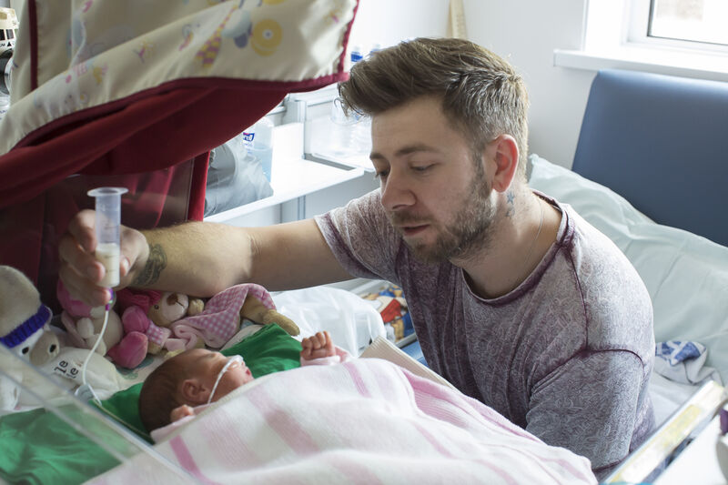 Dad looking over his sleeping baby while holding a syringe of milk above the baby to allow tube feeding