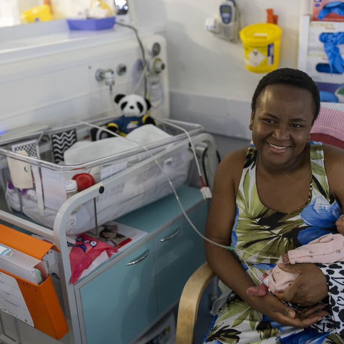 Mum smiling while holding her baby sat next to an incubator
