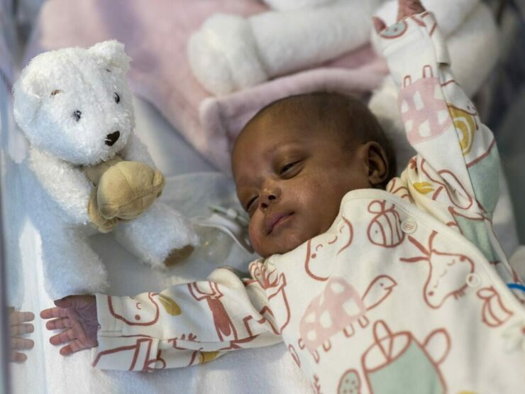 Darker skinned baby lying on back with arms open, next to a teddy bear soft toy