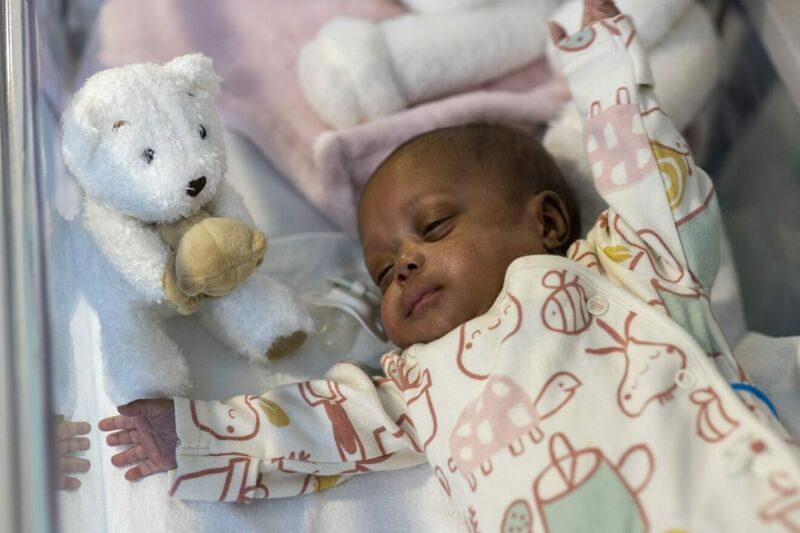 Darker skinned baby lying on back with arms open, next to a teddy bear soft toy