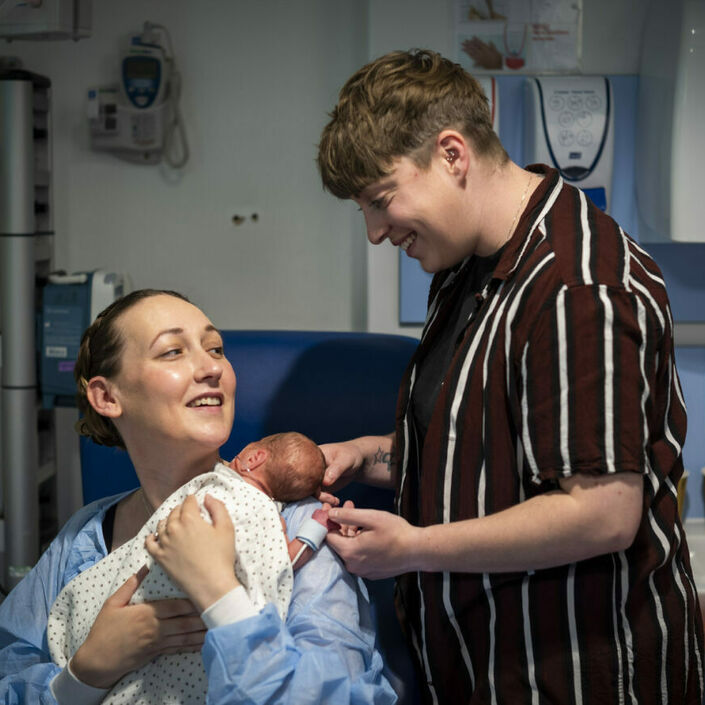 A smiling mum is seated holding their baby. The second smiling mum is standing next to them, holding their baby's hand.