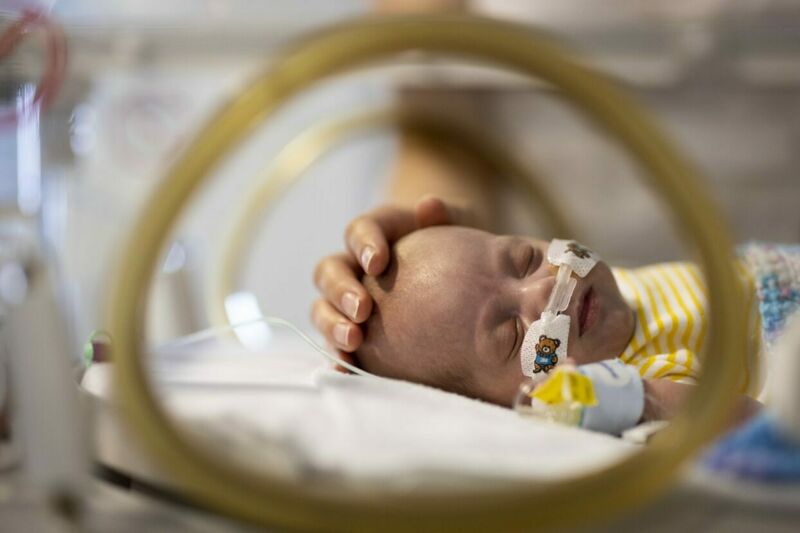 A closeup of a sleeping baby's upper body and head lying in an incubator, with a parent's hand gently placed on their head
