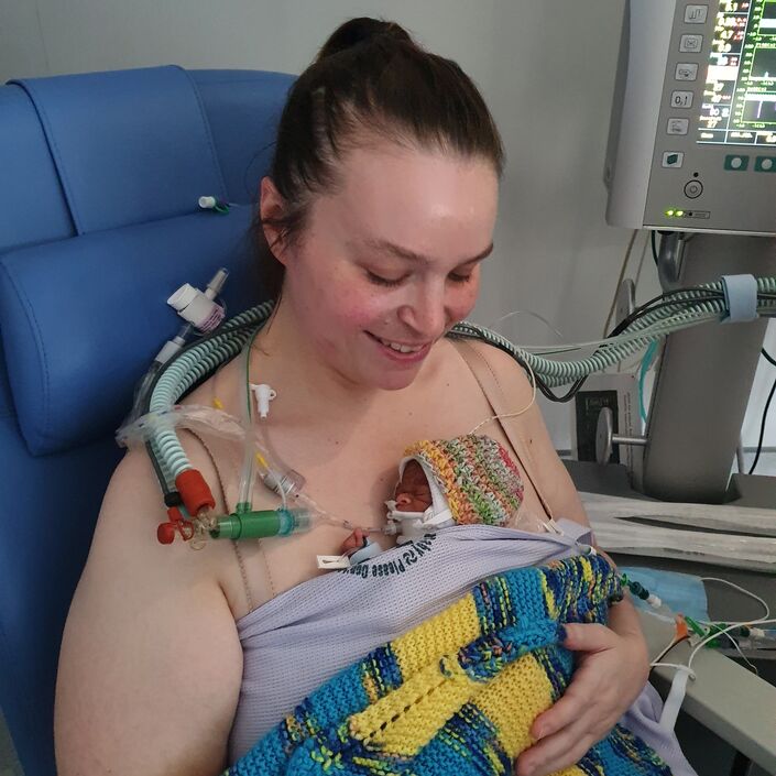 A parent in a hospital chair smiles looking down at their premature baby, who is directly against the skin on their chest, wrapped in a blanket. The baby has some medical tubes and wires attached to them.